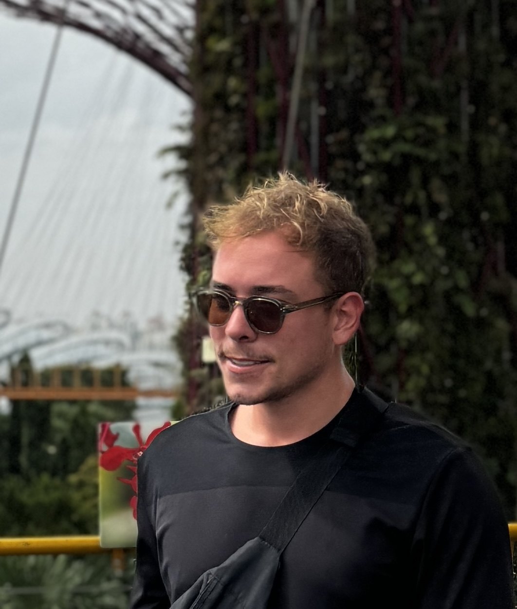 Vitor Magnos, de camiseta preta e óculos escuros, em pé diante de uma parede de vegetação.